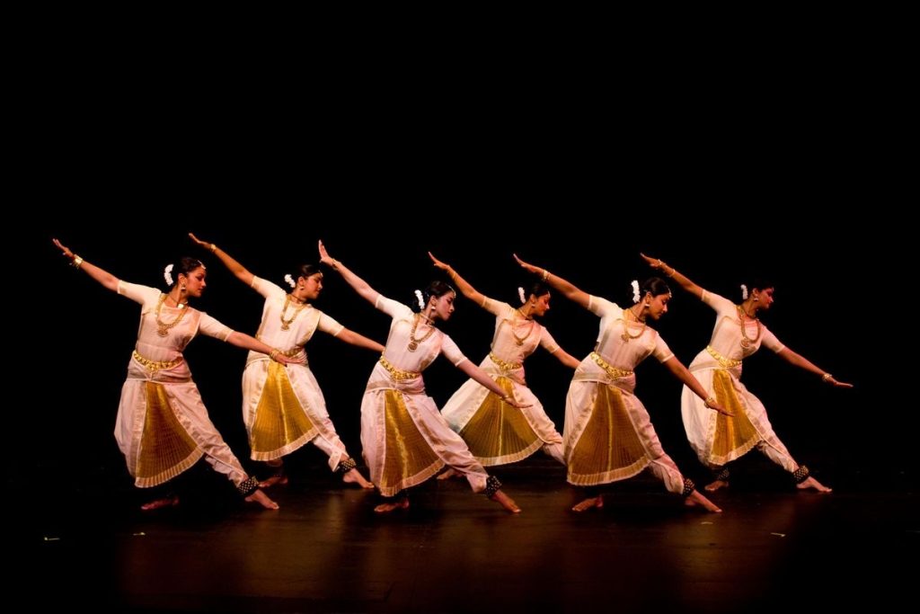 Six young girls gracefully perform a synchronised dance step, dressed in a white classical costume that showcase the beauty of their routine.